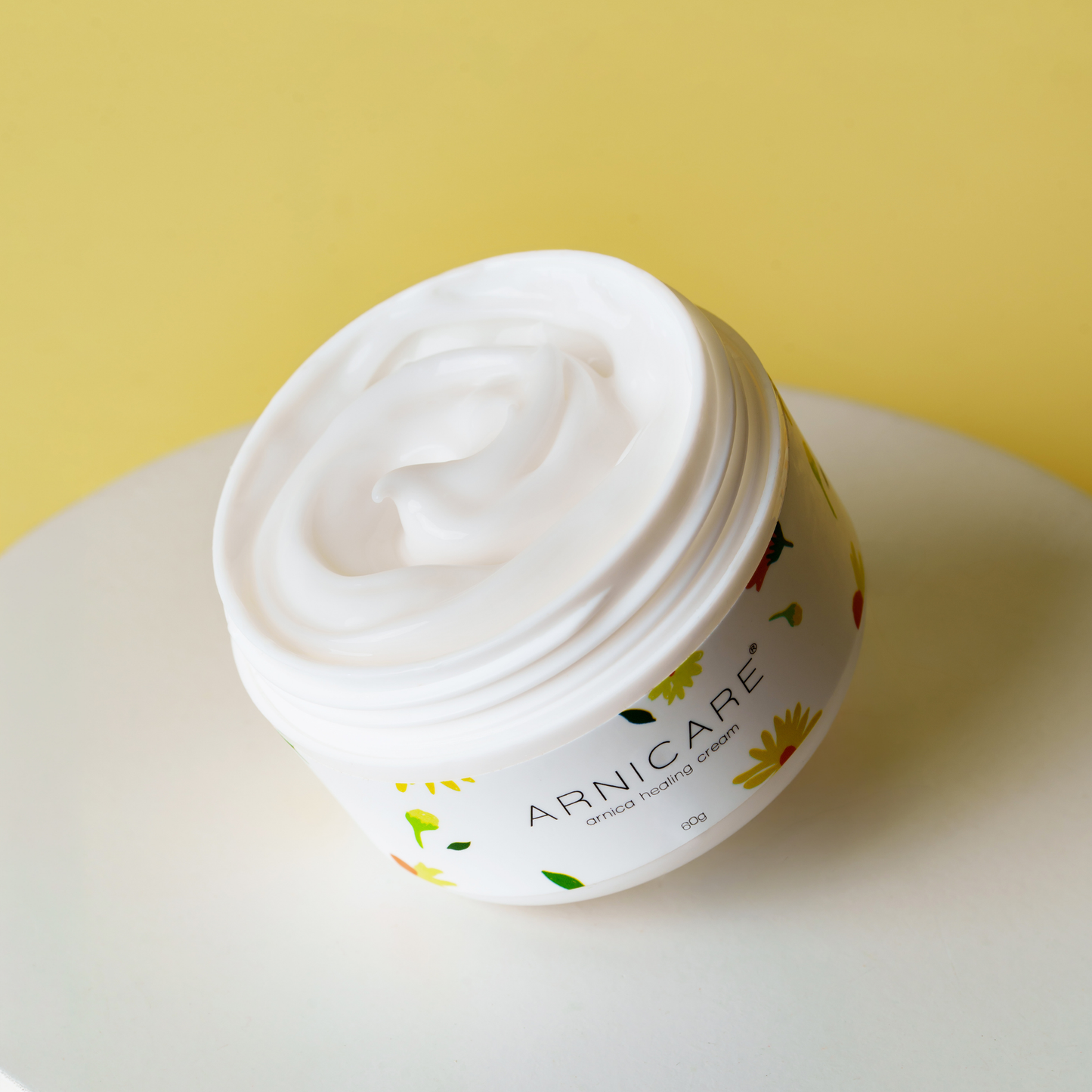 A white jar of ARNICARE healing cream filled with thick white cream, set on a white round surface against a yellow background.
