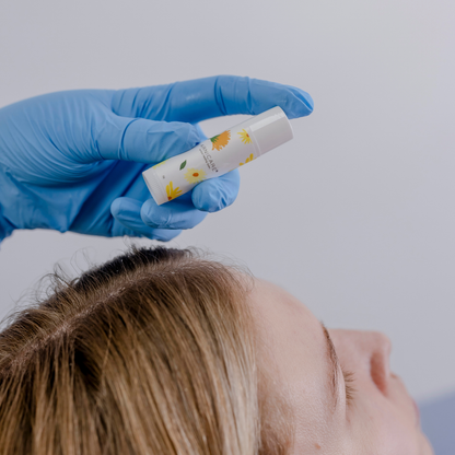 A close-up of a person's head and hair while a gloved hand holds a small white Arnicare Healing Balm with yellow floral graphics above the forehead.