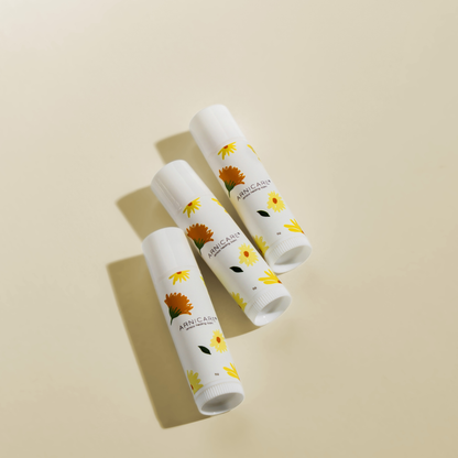 Three white lip balm tubes with a sunflower and daisy floral pattern, arranged diagonally on a warm beige background.