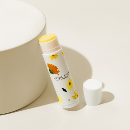 A tube of Arnica Healing balm with a white label decorated with yellow and orange flowers, shown with its cap off next to a beige cylindrical pedestal on a white surface.