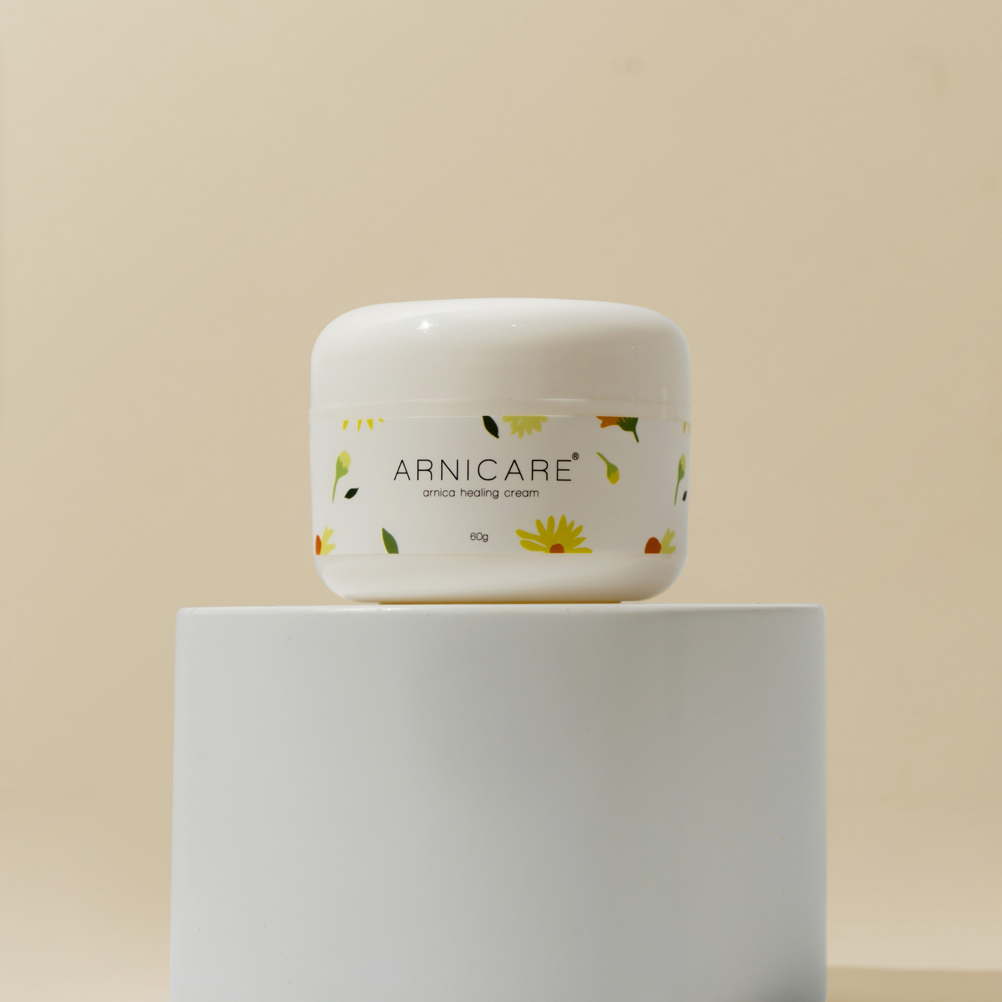 A white plastic jar of ARNICARE arnica healing cream with a floral label, displayed on a white cylindrical pedestal against a warm beige background.