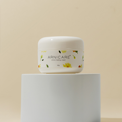 A white plastic jar of ARNICARE arnica healing cream with a floral label, displayed on a white cylindrical pedestal against a warm beige background.