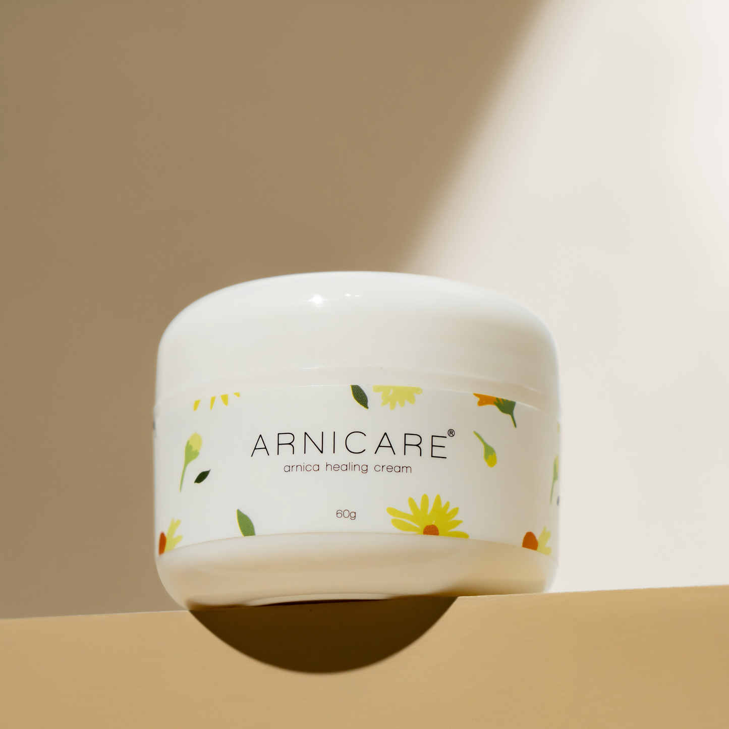A white jar of ARNICARE arnica healing cream with a floral pattern label, set against a warm beige background with a diagonal light beam.