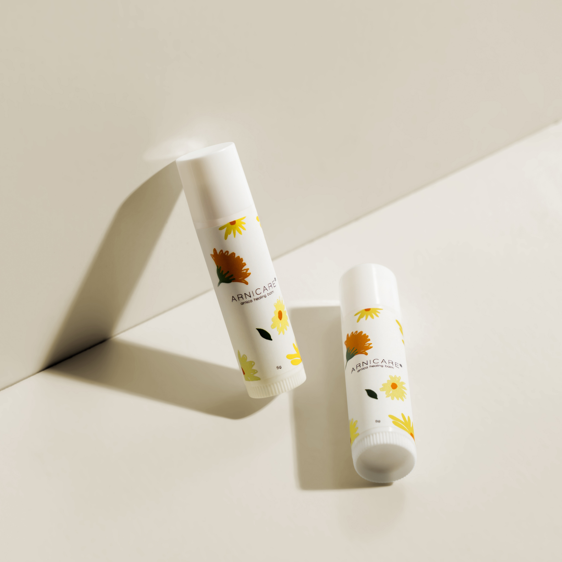 Two white lip balm tubes with yellow and orange floral illustrations, labeled ARNICARE, resting on a clean, light beige surface with soft shadows.