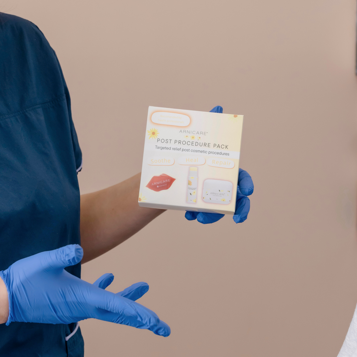 A person wearing blue medical gloves holds a box labeled 'ARNICARE POST PROCEDURE PACK' against a plain beige background.