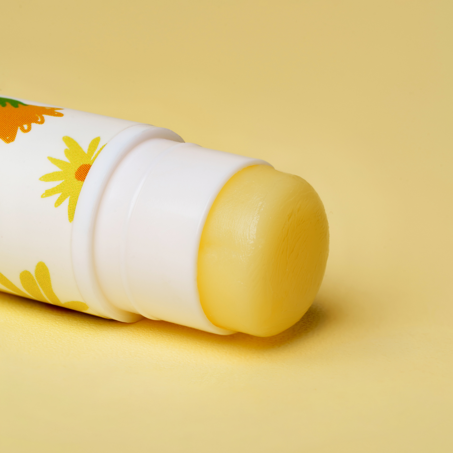 A close-up of a lip balm stick with its white cap removed, showing a smooth yellow balm bar. The tube is white with a yellow and orange daisy pattern, set against a solid pale yellow background.