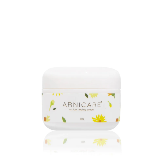 A white plastic jar of ARNICARE arnica healing cream with a white lid, decorated with small yellow flowers and green leaves, set against a solid white background with a soft reflection below.