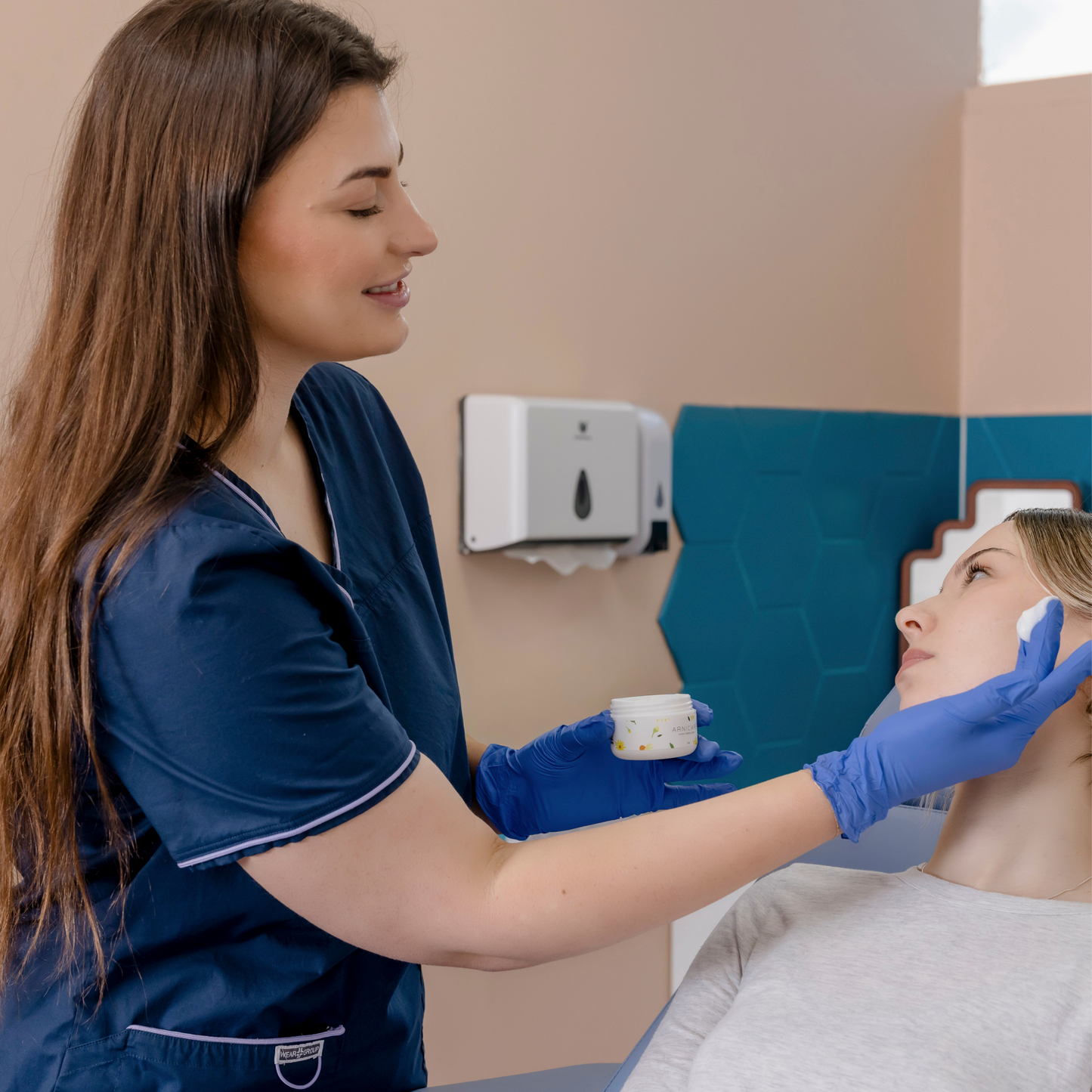 A cosmetic nurse in navy blue scrubs and blue gloves applies a small amount of Arnicare Healing cream to a patients face in a treatment room.