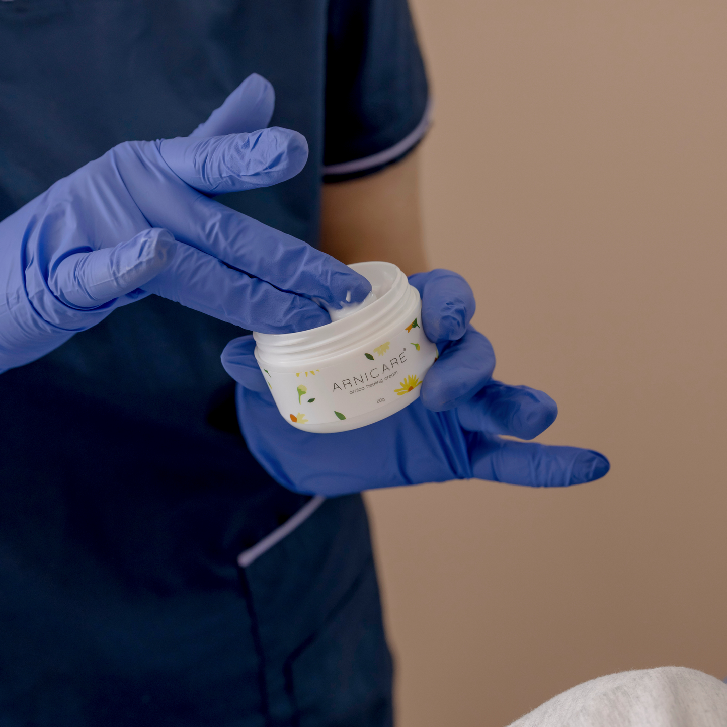 A close-up of a person wearing blue medical gloves holding a small white jar labeled ARNICARE, Healing Cream with the lid being lifted, against a soft beige background.