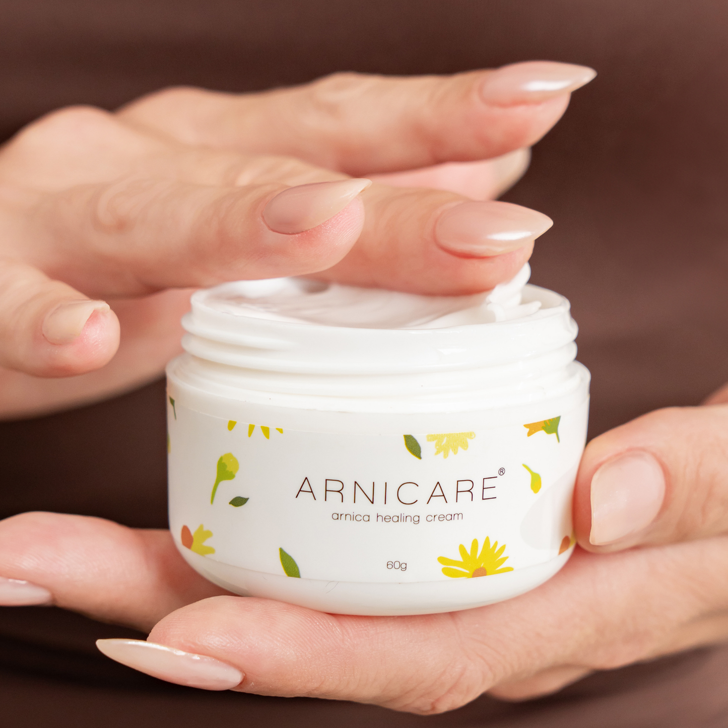A close-up of a person's hands holding a white jar of ARNICARE arnica healing cream, with the cream being spread onto their hand.
