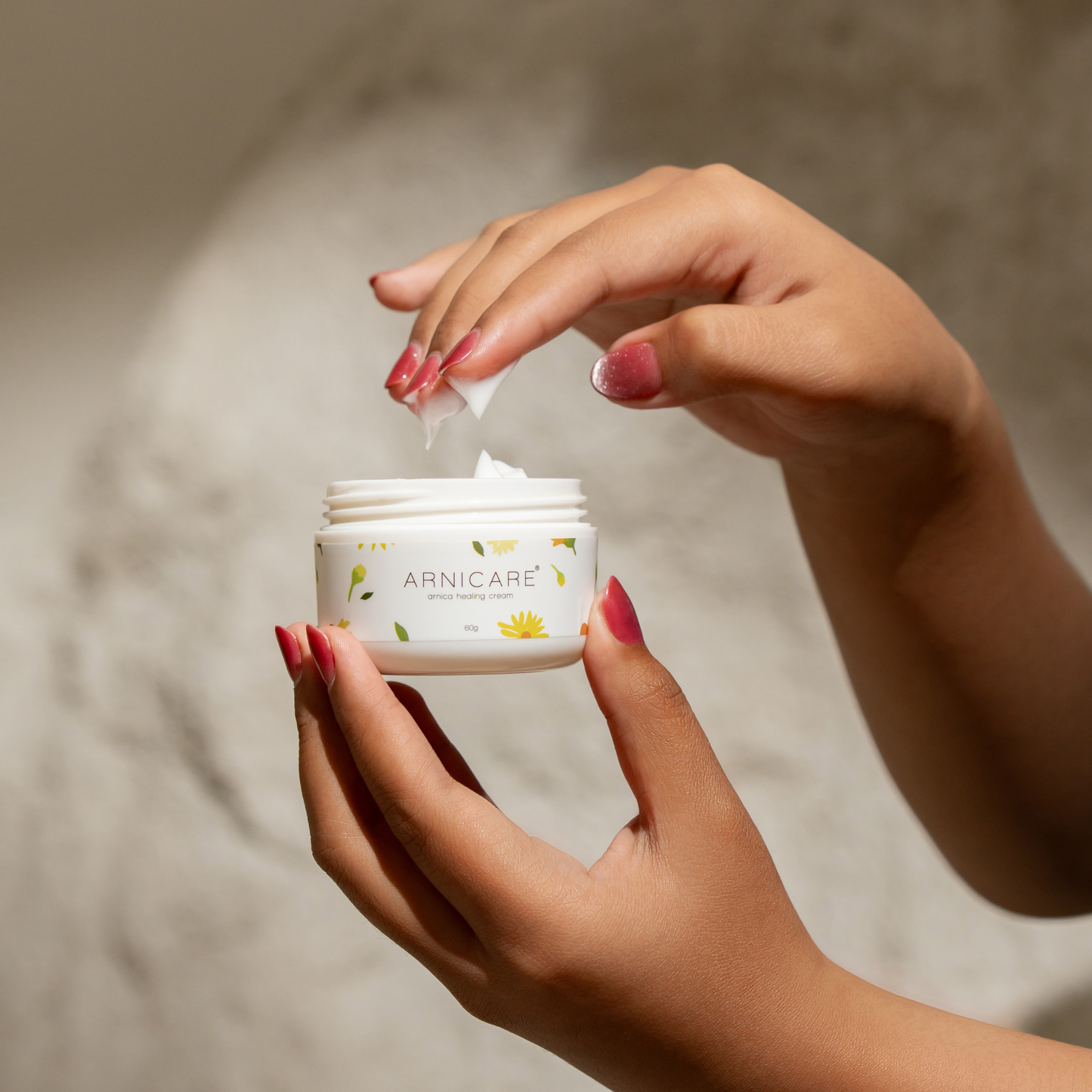 A close-up of a person's hands with pink manicured nails holding an open white jar of Arnicare white healing brusie cream, with a dollop of white cream being spread onto a hand.