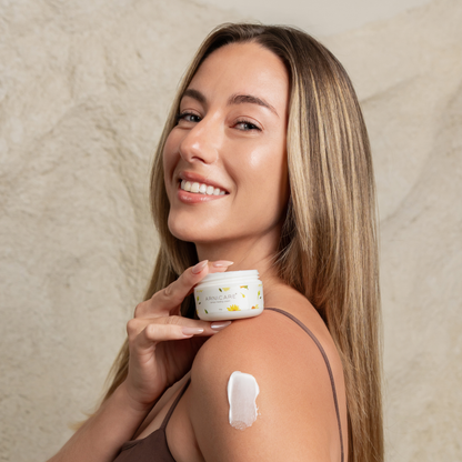 A smiling woman with long blonde hair holds a small white Arnicare Healing Cream near her shoulder, with a dollop of white cream on her upper arm. She is posed against a textured beige stone wall.