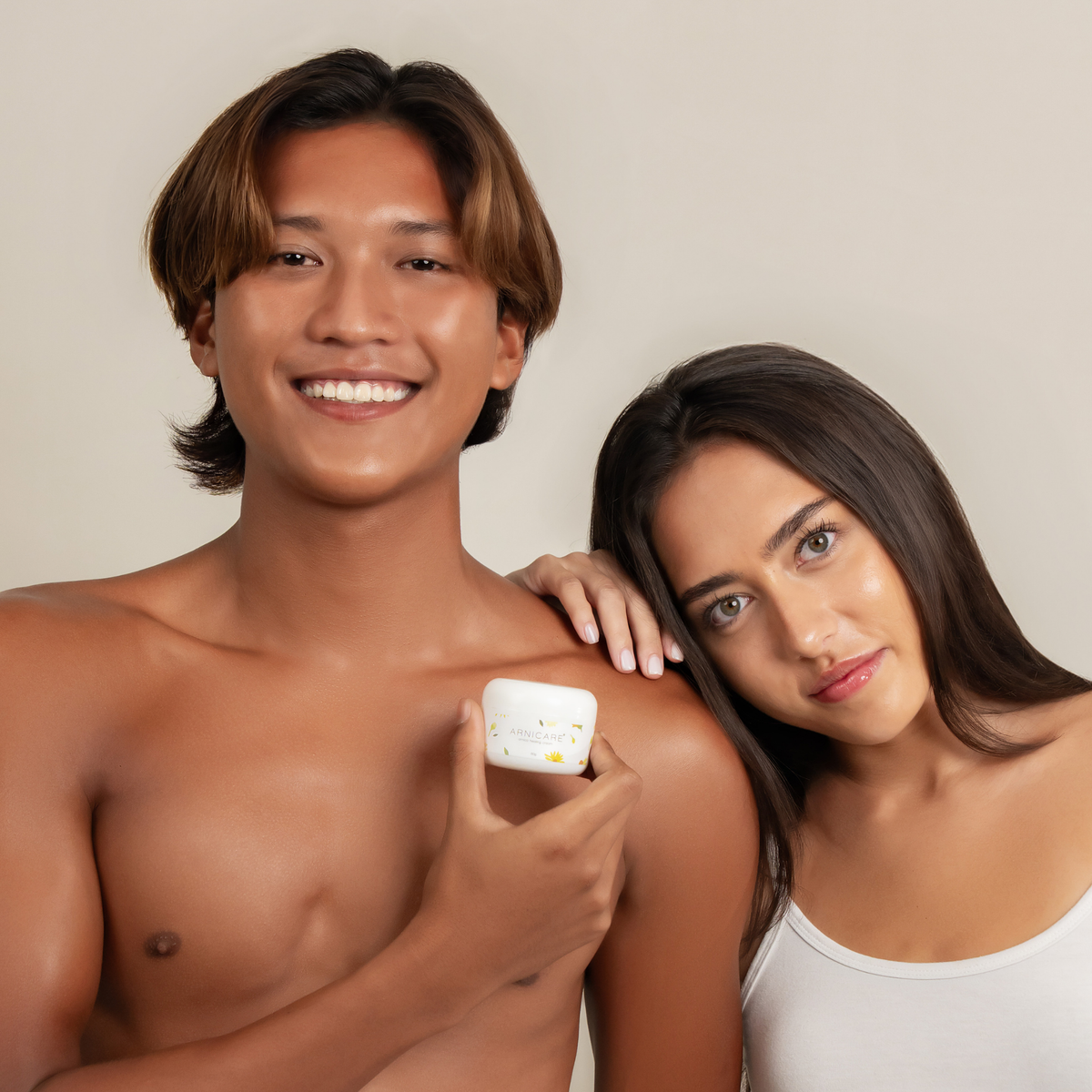 A smiling shirtless man and a woman in a white tank top pose together against a plain light beige background, with the man holding a small white cosmetic jar of Arnicare Healing Cream near his shoulder.