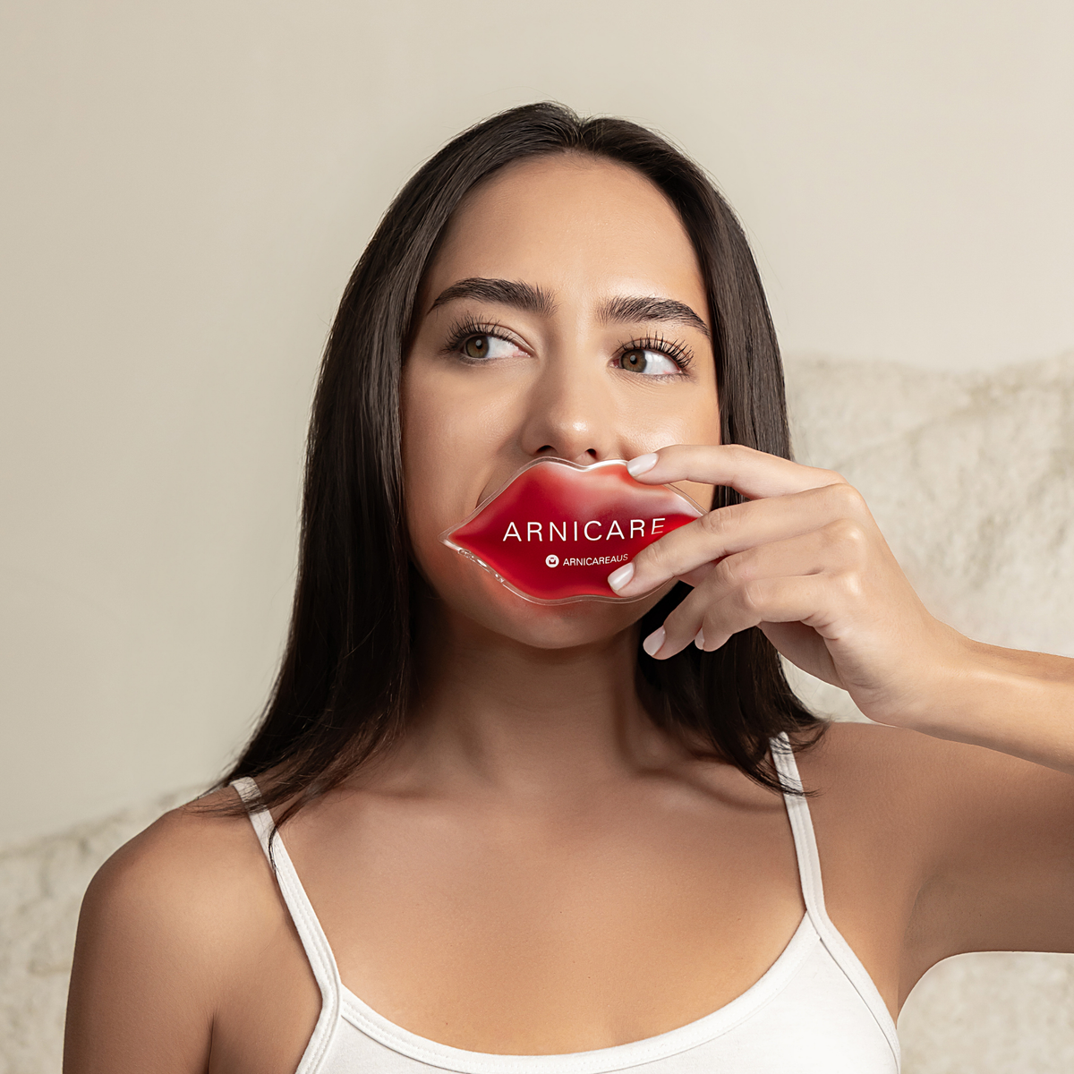 A woman holding a red ARNICARE lip-shaped product to her lips, wearing a white camisole against a soft beige background.
