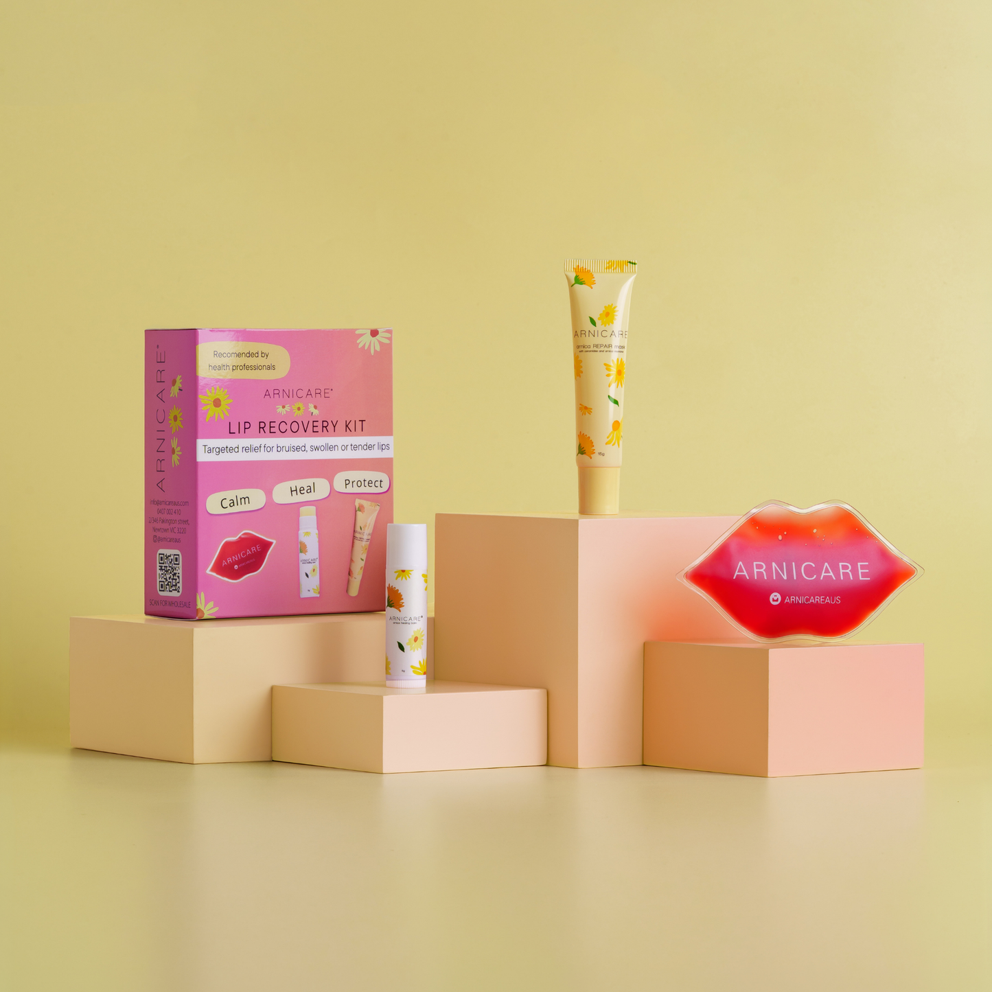 Arnicare Lip Recovery Kit displayed on peach-colored pedestals against a soft yellow background. The kit includes a pink product box, a yellow and white tube, a small white tube, and a red lip-shaped item with the Arnicare logo.