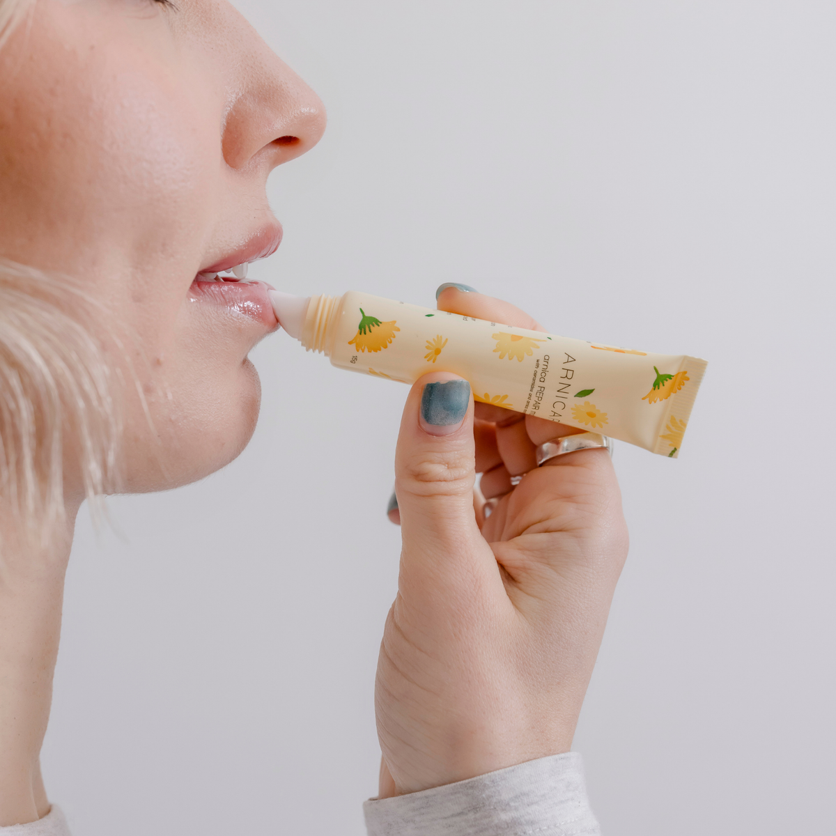 A close-up of a person applying Arnicare Lip Repair Mask from a small tube with a floral print. The tube is cream-colored with yellow flowers and green leaves, and the brand name 'ARNICA' is visible on the packaging.