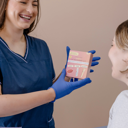 A smiling woman in blue medical scrubs and blue gloves holds a pink box labeled 'Lip Recovery Kit' while showing it to another woman in a clinical indoor setting.