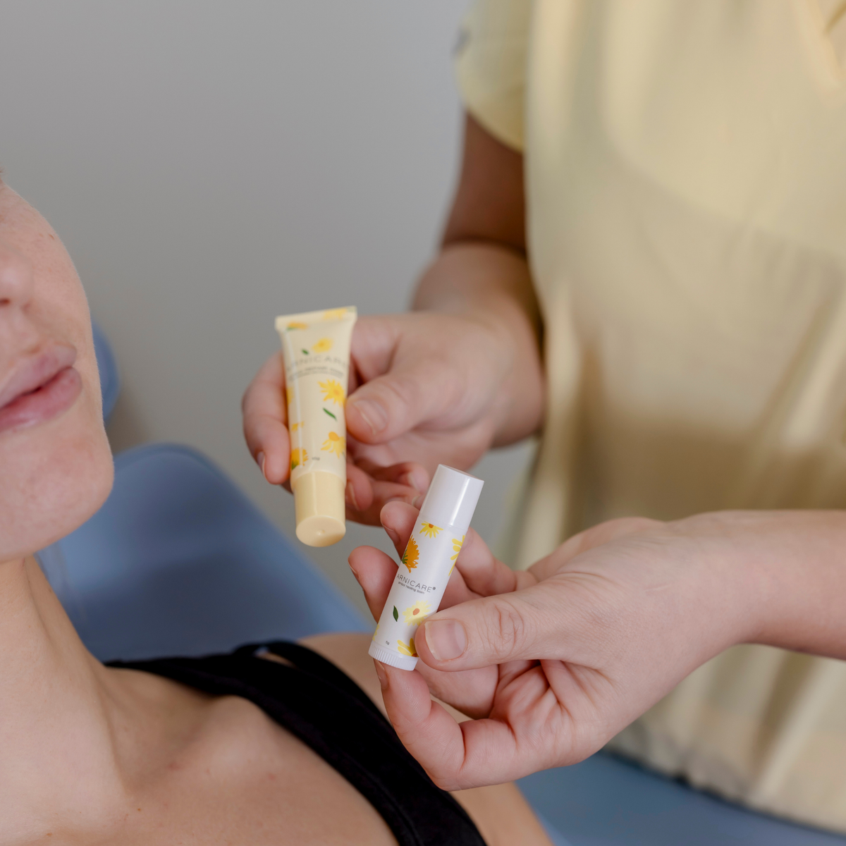 A close-up lifestyle shot of a skincare treatment. A cosmetic nurse in a pale yellow uniform holds two tubes of the product: a larger cream-colored tube with a yellow floral pattern and a smaller white tube with a white cap.