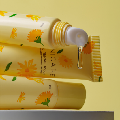 Three yellow tubes of ARNICARE lip repair mask with a daisy floral pattern, one of which is open with a clear gel drop dripping from the nozzle, set against a pale yellow background.