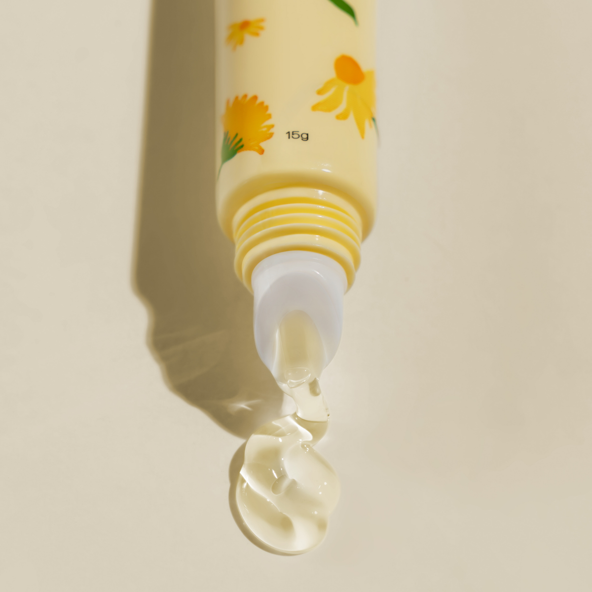 A close-up of a pale yellow Arnicare Lip Repair Mask with yellow flower illustrations, shown with a clear gel-like product dripping from the tip against a warm beige background.