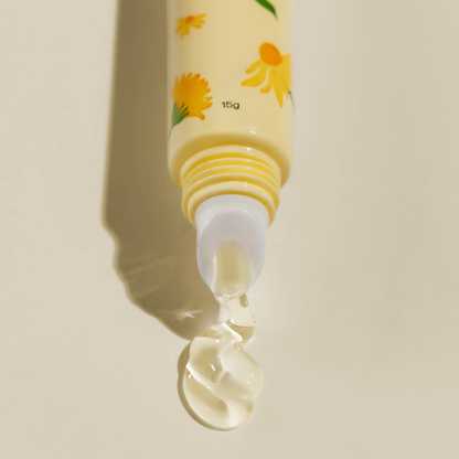 A close-up of a pale yellow Arnicare Lip Repair Mask with yellow flower illustrations, shown with a clear gel-like product dripping from the tip against a warm beige background.