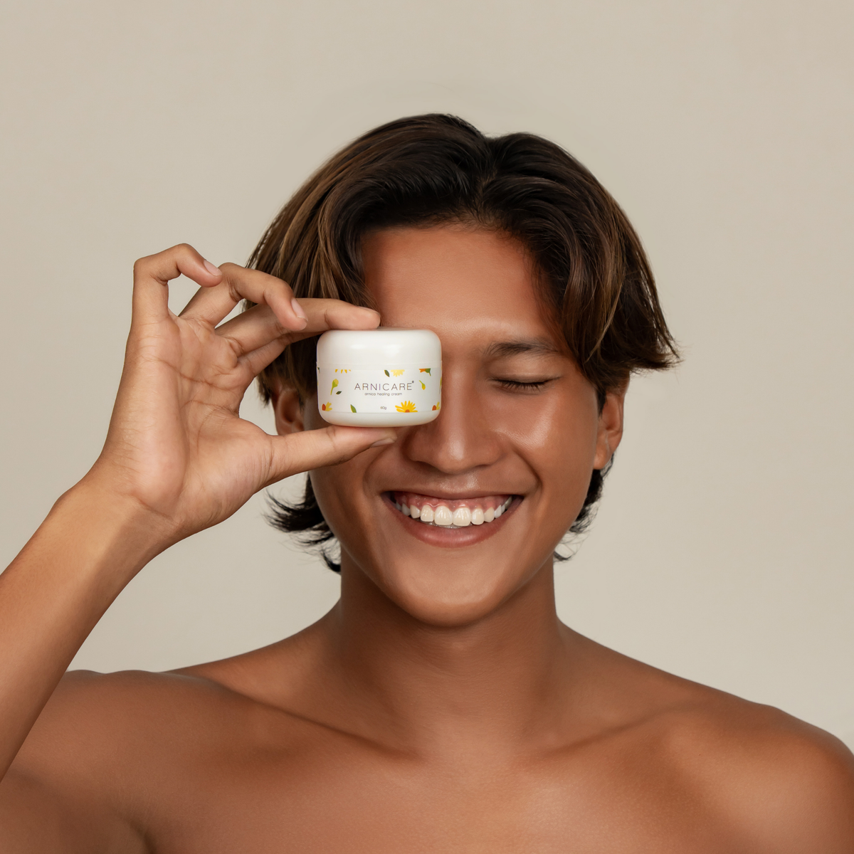 A smiling man with short brown hair holds a small white cosmetic jar of Arnicare Healing Cream over his eye against a plain warm beige background.