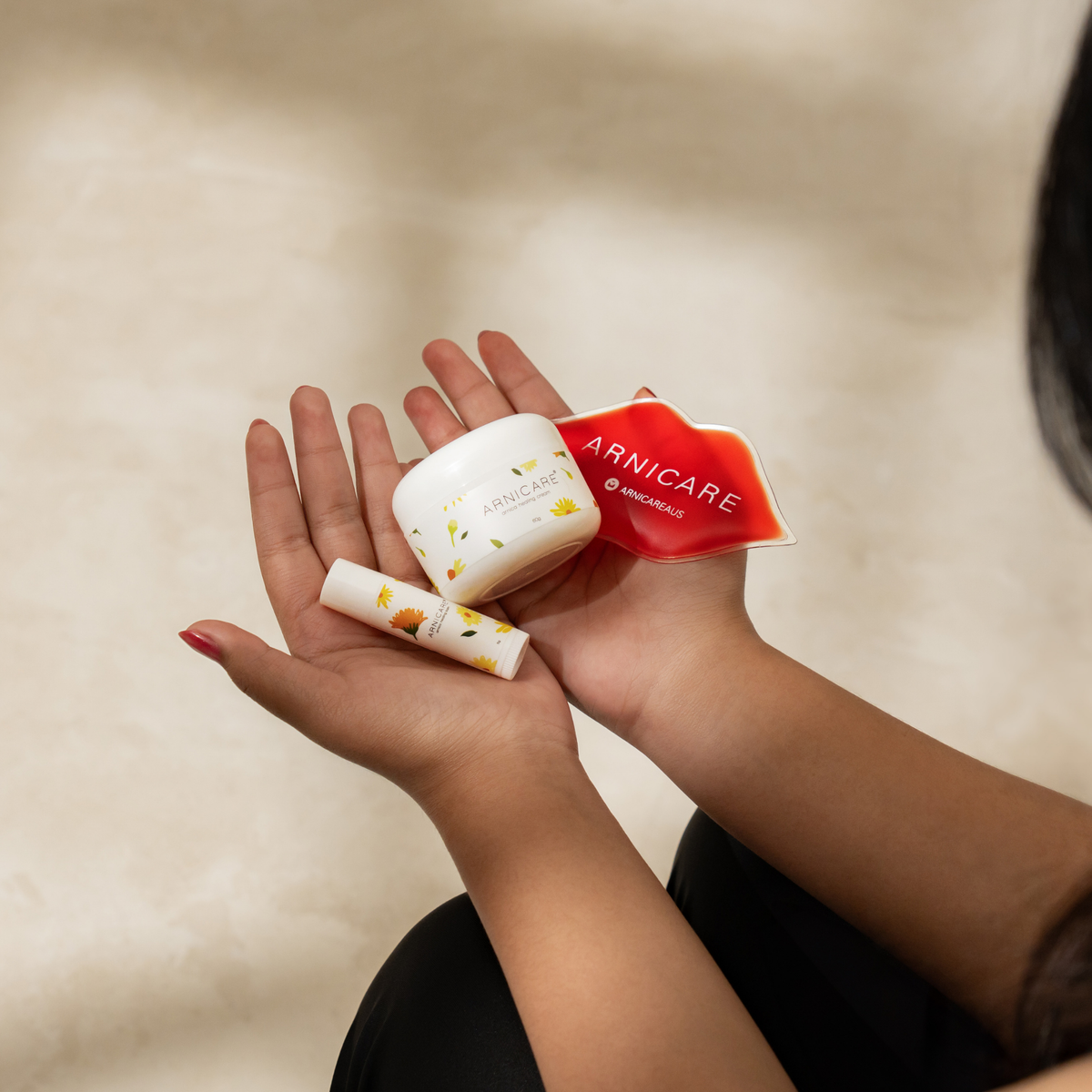 A person holds three Arnicare products in their hands against a warm beige background: a white jar of Arnicare Healing cream, a matching Arnicare Healing Balm, and a red heart-shaped Arnicare lip shaped ice pack.