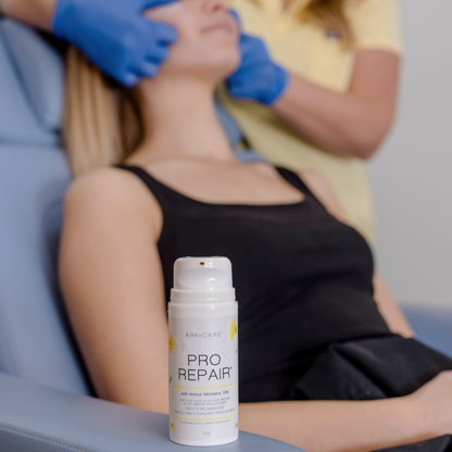 A white and yellow Arnicare Pro Repair ampoule bottle is shown in the foreground on a light blue treatment chair, with a woman receiving a facial treatment in the blurred background.