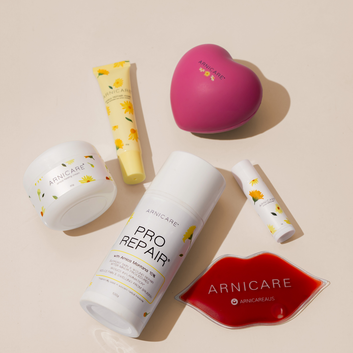 A flat-lay arrangement of five Arnicare skincare products on a warm beige background, including a Arnicare Healing Cream, a yellow Arnicare Repair Lip Mask a white Arnicare Pro Repair bottle, a pink heart-shaped stress ball item, a small white Arnicare Healing Balm and a red lip-shaped ice pack.