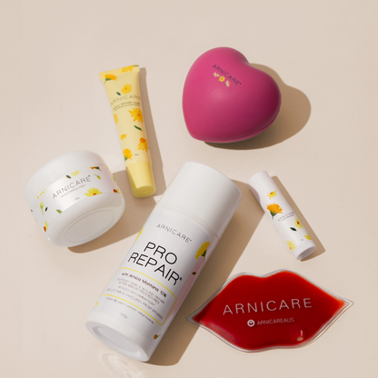 A flat-lay arrangement of five Arnicare skincare products on a warm beige background, including a Arnicare Healing Cream, a yellow Arnicare Repair Lip Mask a white Arnicare Pro Repair bottle, a pink heart-shaped stress ball item, a small white Arnicare Healing Balm and a red lip-shaped ice pack.