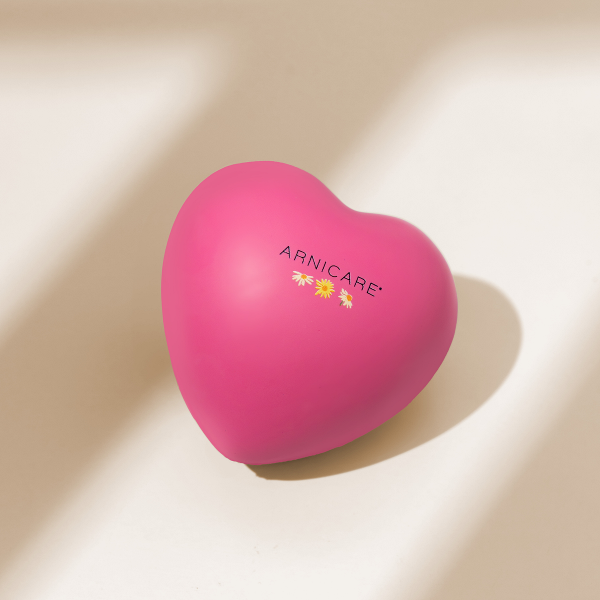A heart-shaped pink Arnicare stress ball with a small daisy and sunflower graphic and the brand name printed on the front, resting on a warm beige surface with soft diagonal shadows.
