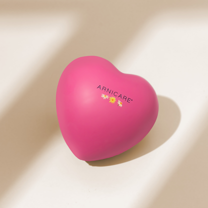 A heart-shaped pink Arnicare stress ball with a small daisy and sunflower graphic and the brand name printed on the front, resting on a warm beige surface with soft diagonal shadows.